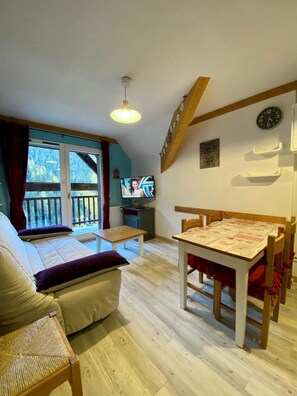 Een gezellige kamer met een bed, een houten tafel, een televisie en een balkon met uitzicht op bomen.