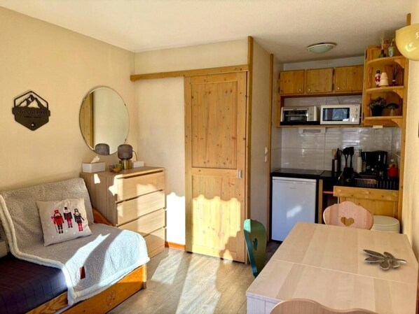 Una habitación compacta con cama, una mesita pequeña, kitchenette y un armario de madera.