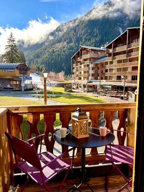 Un balcón de madera con una mesa y sillas, con vistas a un paisaje montañoso donde se observa un edificio de hotel al fondo.