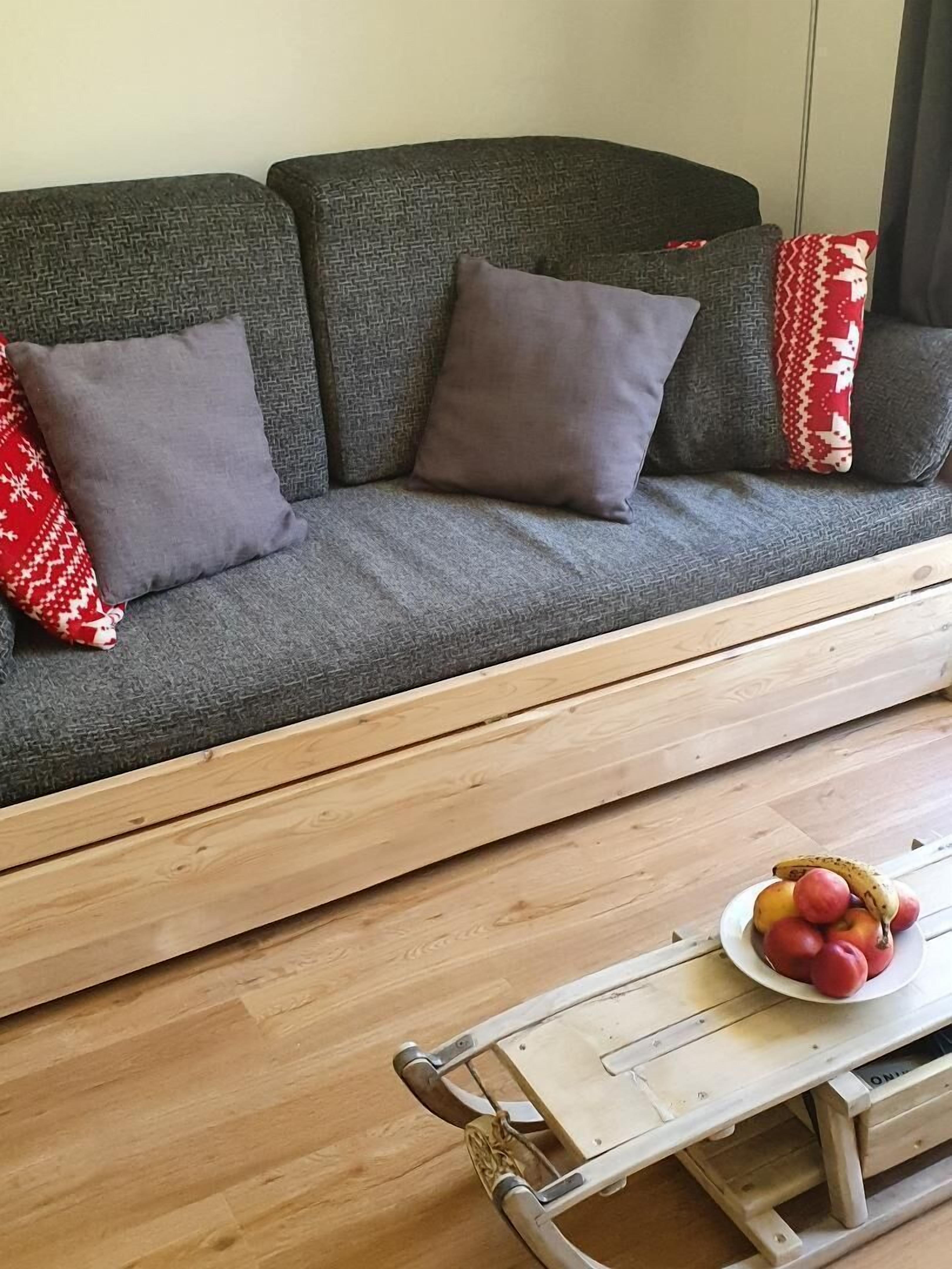 Een grijze bank met drie kussens, een houten salontafel met een fruitschaal en een houten vloer.