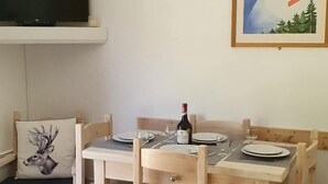 Uma mesa de jantar de madeira posta com pratos, copos e uma garrafa de vinho. Uma televisão está fixada na parede acima da mesa.