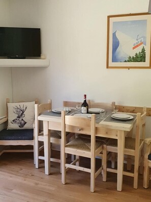 Uma mesa de jantar de madeira posta com pratos, copos e uma garrafa de vinho. Uma televisão está fixada na parede acima da mesa.