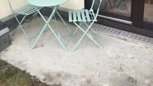 Uma pequena área externa com espaço para sentar, incluindo uma mesa redonda e duas cadeiras, ao lado de uma porta de vidro deslizante.