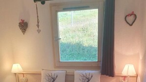 Quarto com cama, duas mesas de cabeceira com abajures, janela com vista para o verde e decorações nas paredes.