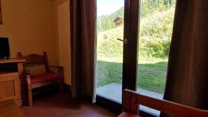 A room with a bed, a desk, and a chair. There is a sliding glass door leading to a grassy outdoor area with trees and a building. - Ski-in/ski-out & foot of the slopes, Ground floor, view mountain, terrace, balcony, tv, ski locker (Valfréjus)
