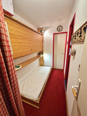 A narrow hotel room with a bunk bed, a red carpet, a wooden wall, a door with a red frame, and a wall clock.