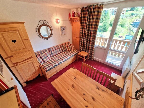 A cozy room with a wooden table, a plaid sofa, a small wooden cabinet, and a window with a view of greenery.