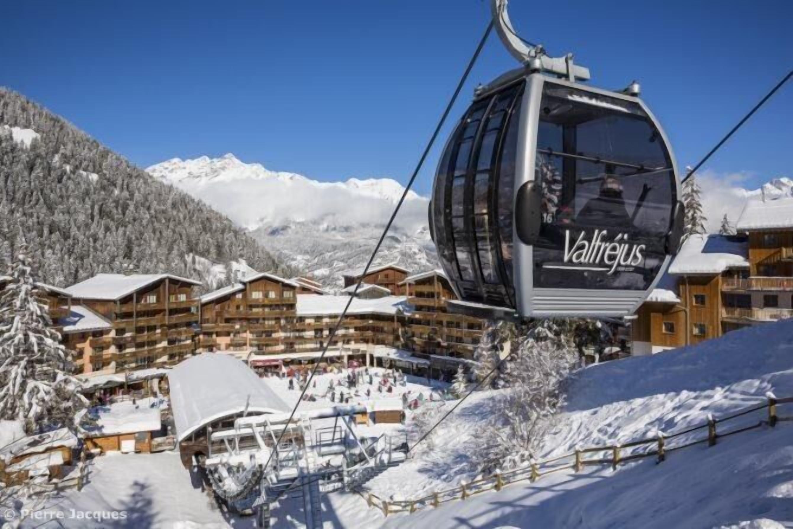 A ski lift with 'Valfréjus' branding, wooden chalets, and a snowy mountain backdrop.