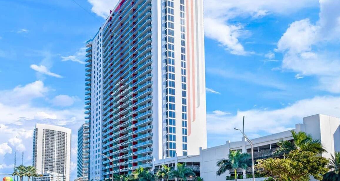 A tall hotel with multiple balconies, surrounded by palm trees and other buildings.