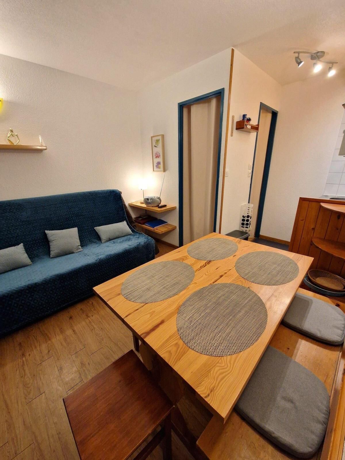 Un espacio habitable compacto con un sofá azul, una mesa de comedor de madera con cuatro individuales tejidos, una pequeña mesita de noche con una lámpara, y una zona de cocina con armarios de madera.