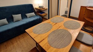 Un espacio habitable compacto con un sofá azul, una mesa de comedor de madera con cuatro individuales tejidos, una pequeña mesita de noche con una lámpara, y una zona de cocina con armarios de madera.