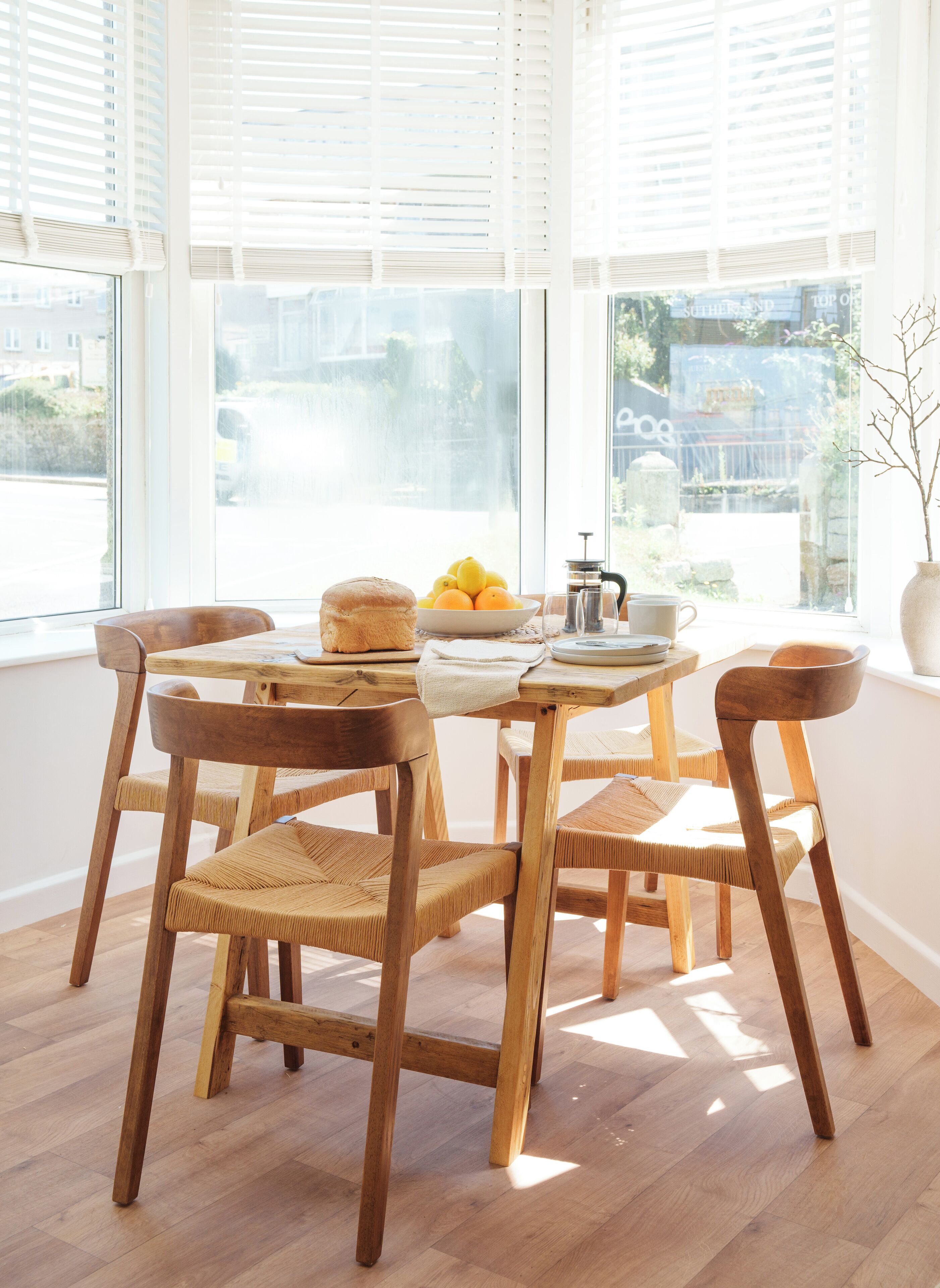 Un comedor con sillas de madera y una mesa, un frutero con fruta y una cafetera.