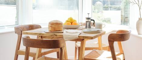 Un comedor con sillas de madera y una mesa, un frutero con fruta y una cafetera.