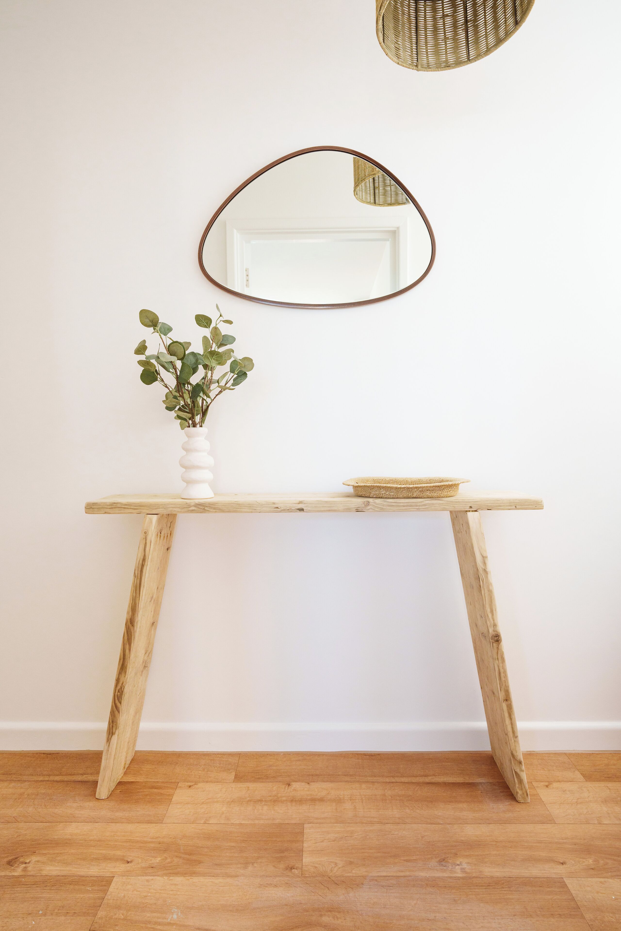 Une table en bois sur laquelle sont posés un vase blanc et un bol, le tout adossé à un mur uni, avec un miroir rond au-dessus.