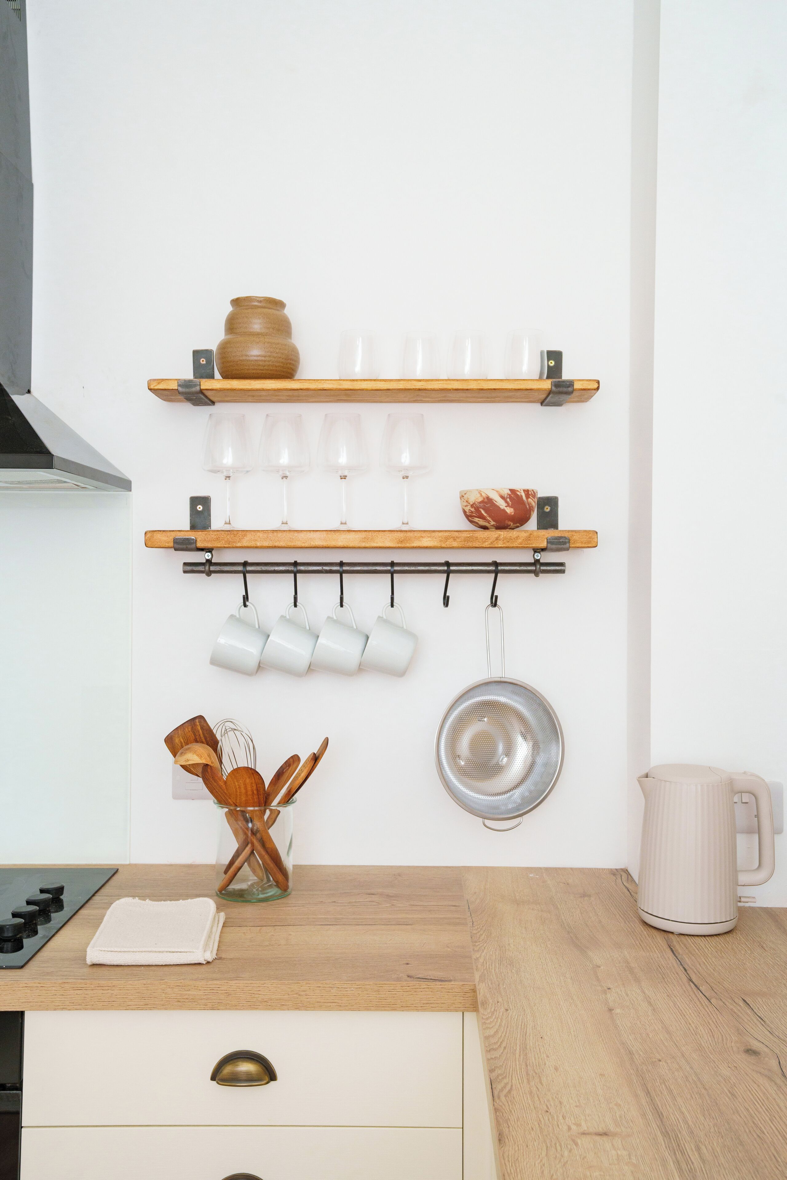 A kitchen with wooden shelves holding a vase, glasses, and a pot. There are hanging mugs and a strainer on the wall. A kettle and a knife block are on the counter.