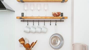 Une cuisine avec des étagères en bois où sont posés un vase, des verres et un pot. Des tasses sont accrochées au mur, ainsi qu’une passoire. Une bouilloire et un range-couteaux se trouvent sur le plan de travail.
