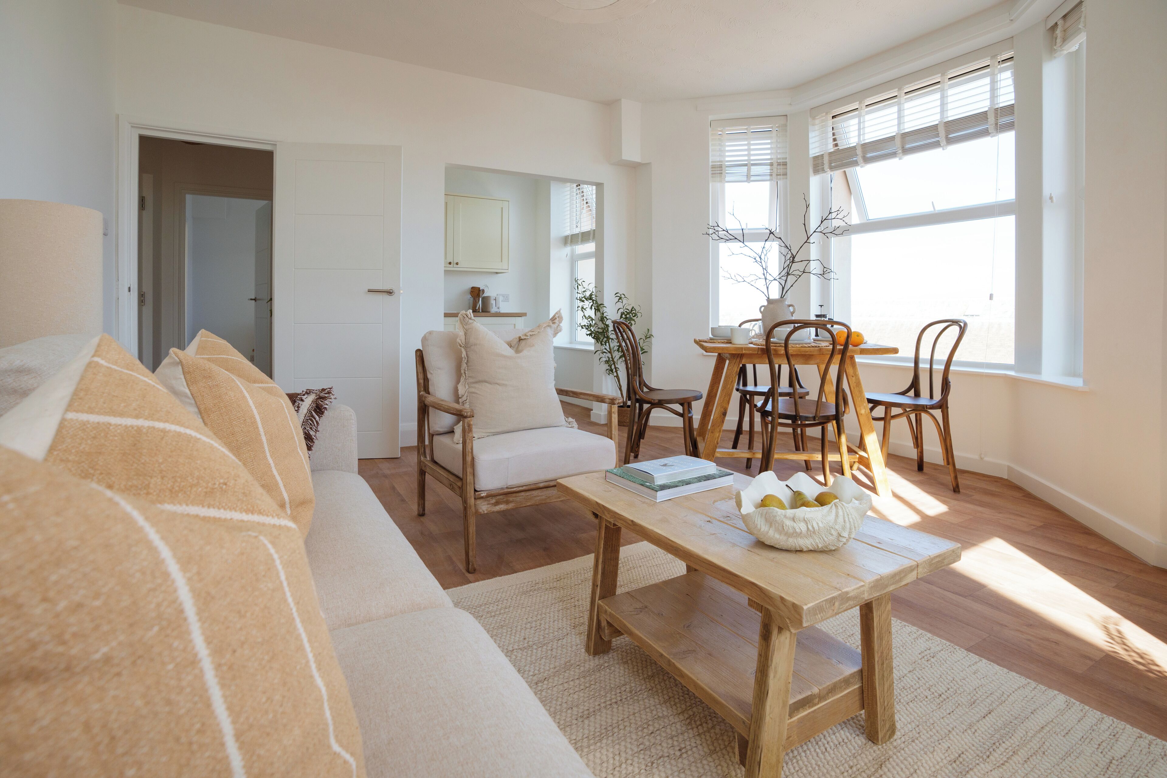 A living room with a sofa, a wooden coffee table with a bowl of fruit, a dining area with a table and chairs, and a large window with blinds.