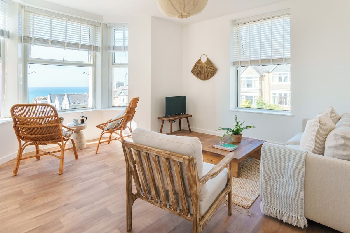 A spacious living room with large windows, a wooden floor, a white sofa, and two wicker chairs.