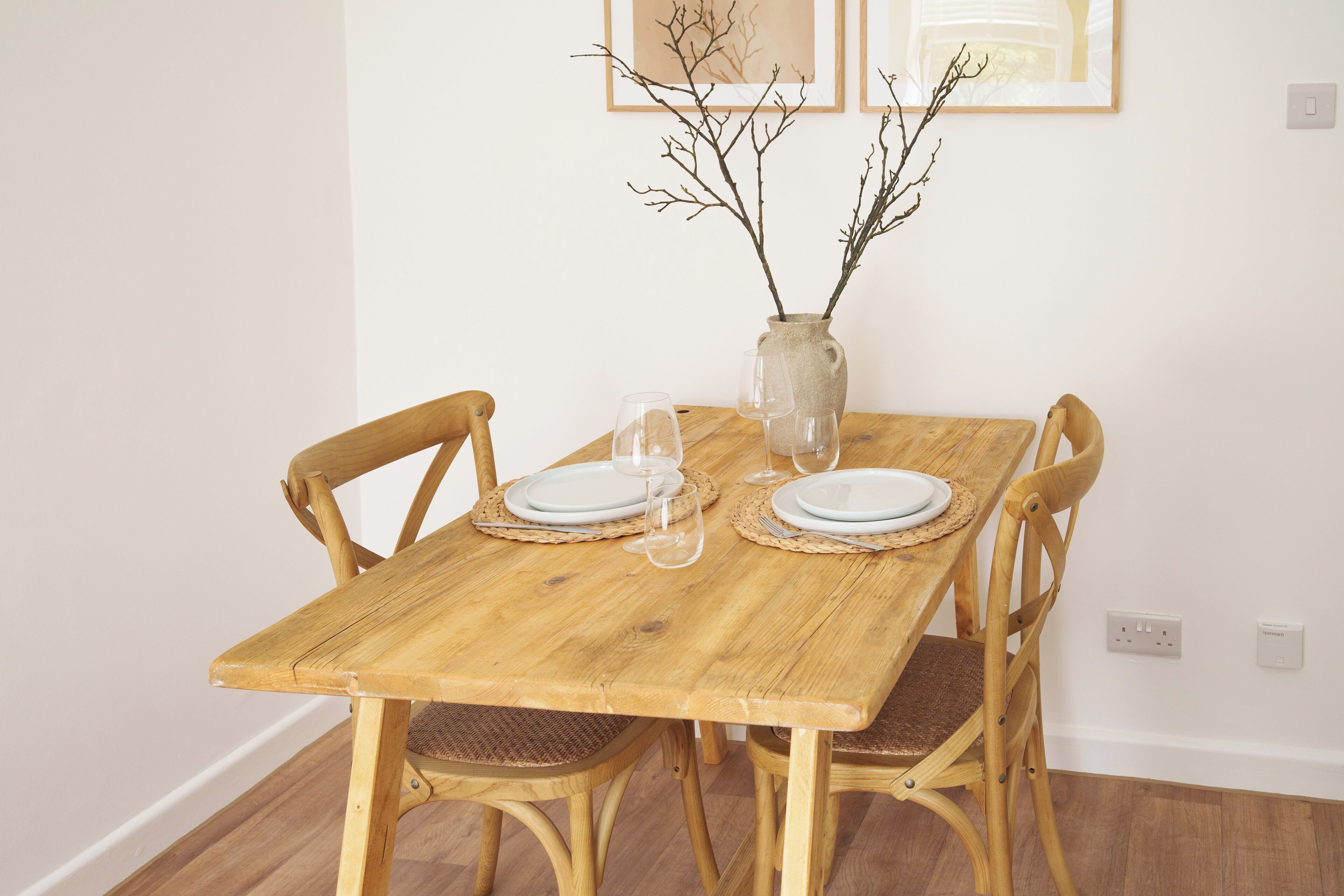 Une table à manger en bois, accompagnée de quatre chaises, un vase décoré de branches et deux œuvres d’art encadrées accrochées au mur.