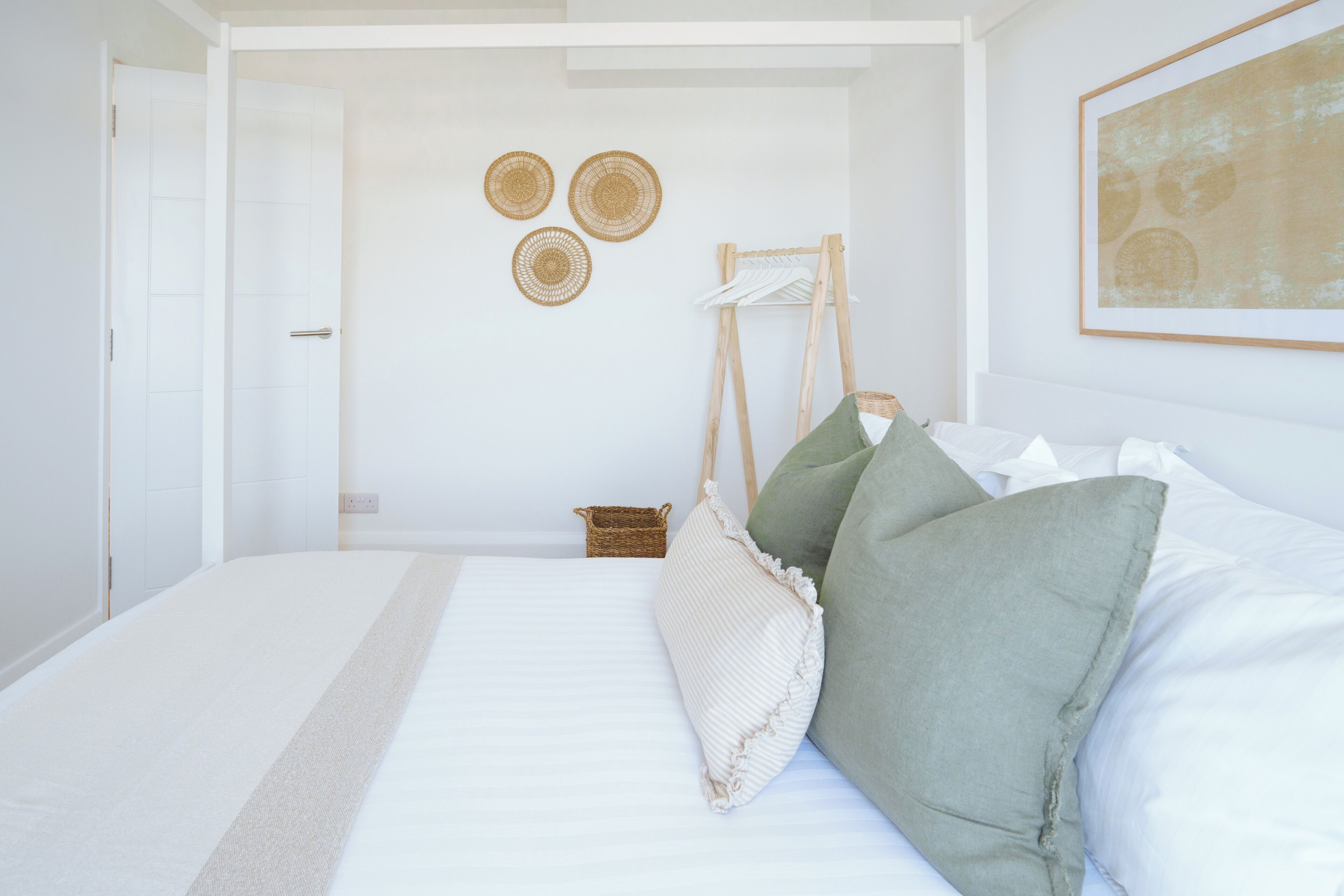 Cama bem arrumada com lençóis brancos e alguns travesseiros, um pequeno cesto de palha e um cavalete de madeira com um pano branco.