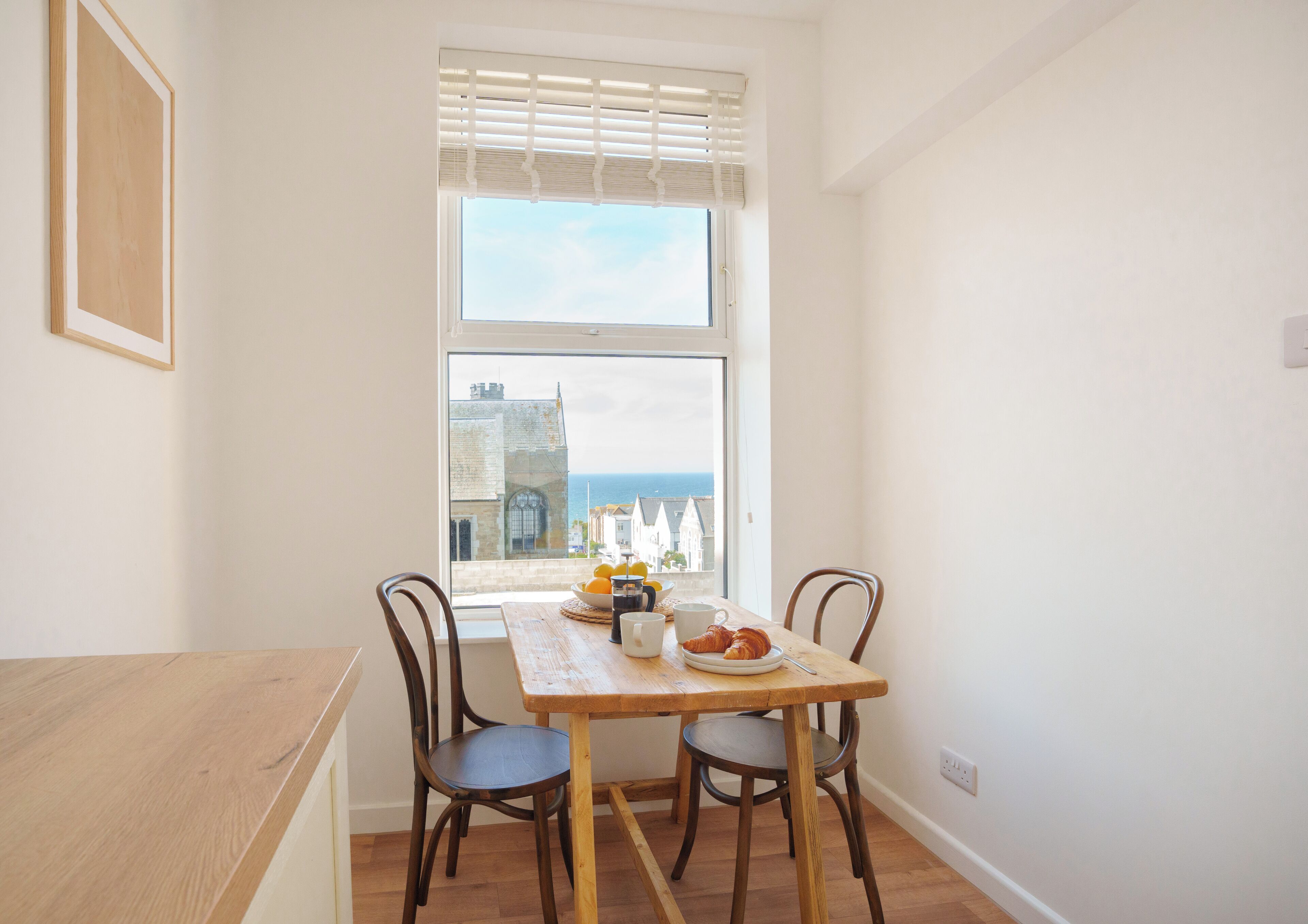 Un coin repas avec une table et des chaises en bois, une fenêtre avec des stores et une vue sur la mer et les bâtiments.