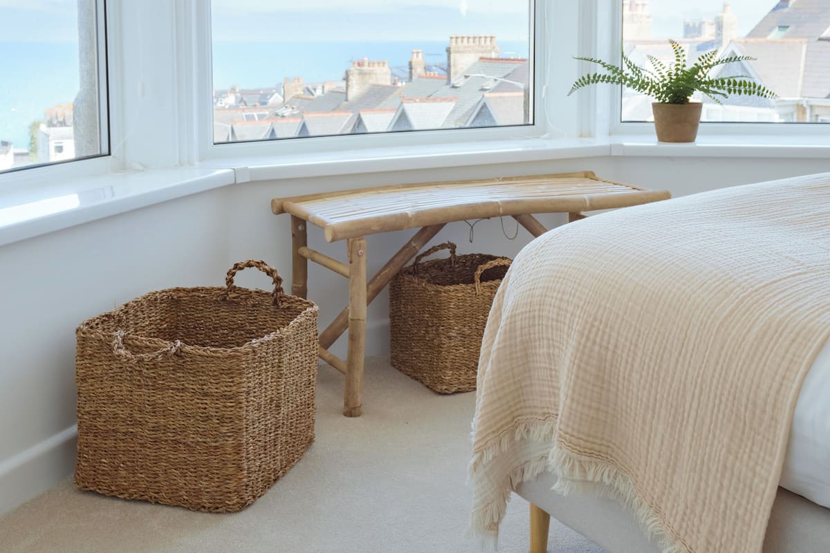 A bedroom with a large window, a bed, two wicker baskets, a small wooden table, and a potted plant.