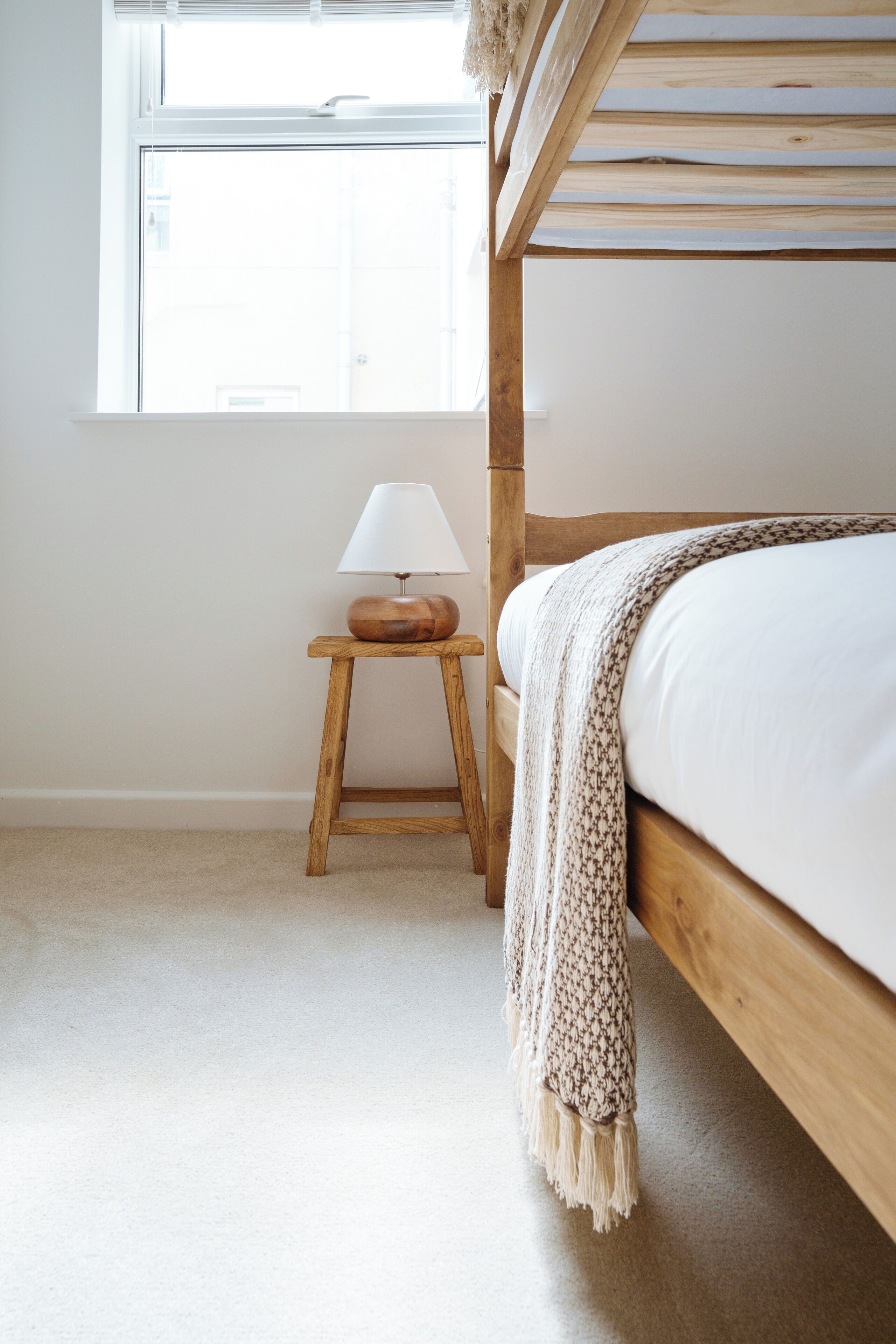 A bunk bed with a wooden frame, a bedside table with a lamp, and a window with a view of the outside.