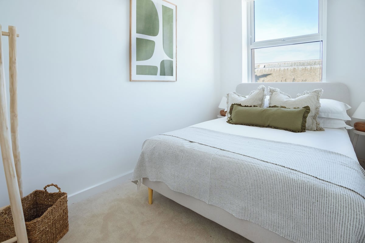 A neatly arranged bedroom with a bed, a bedside table, a lamp, a framed abstract artwork, a window, and a wooden ladder leaning against the wall.