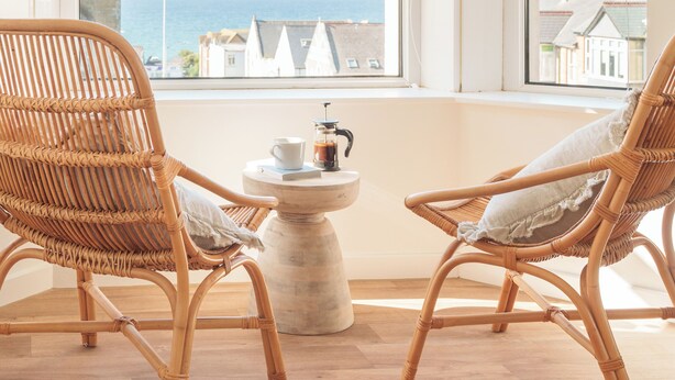 A room with two wicker chairs facing a window overlooking the sea. A small table with a coffee pot and cup is in the center. - VESTO  Coastal Hideout (Newquay)