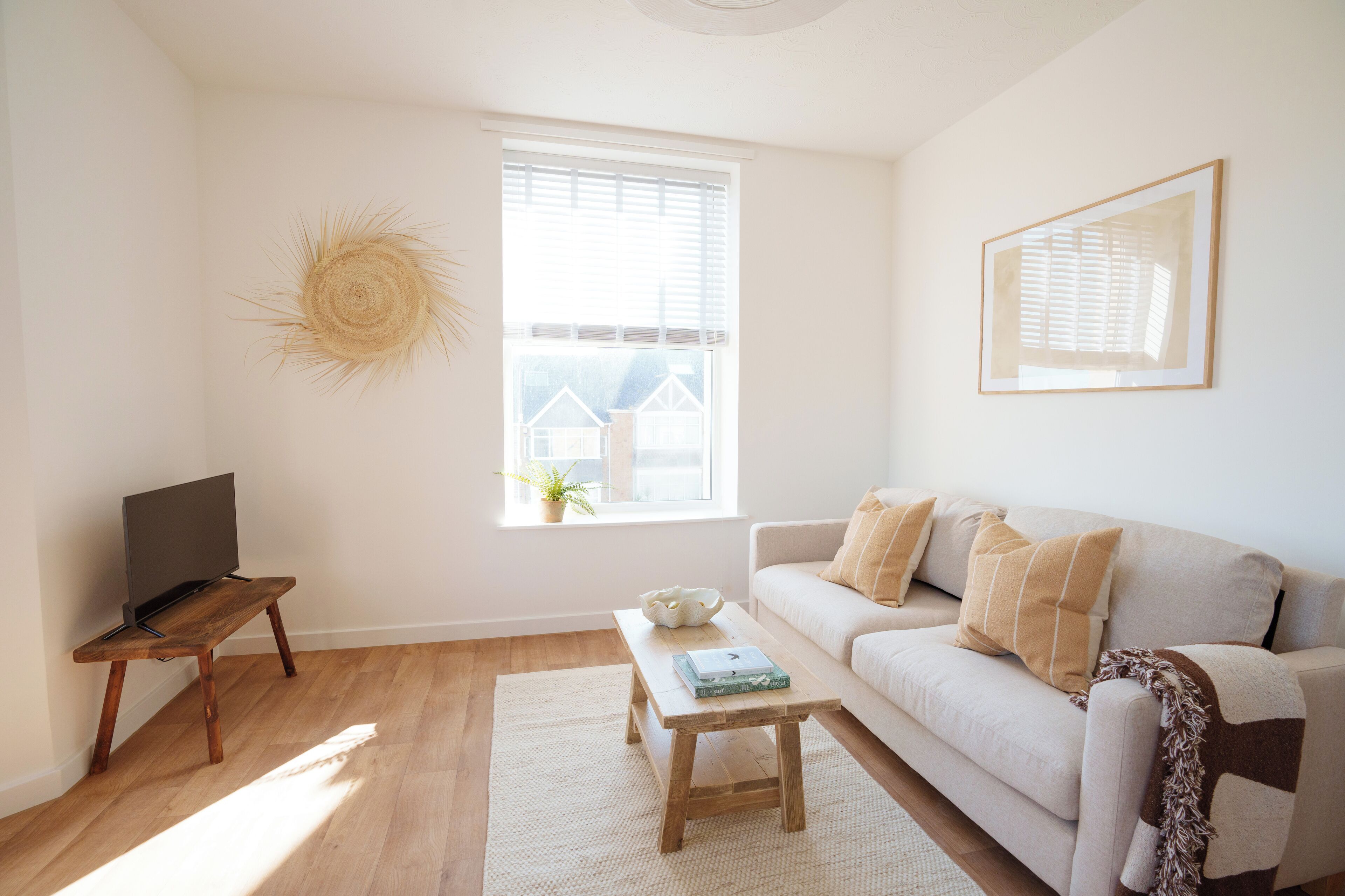 A living room with a wooden coffee table, a white sofa, a TV on a wooden stand, and a window with blinds.