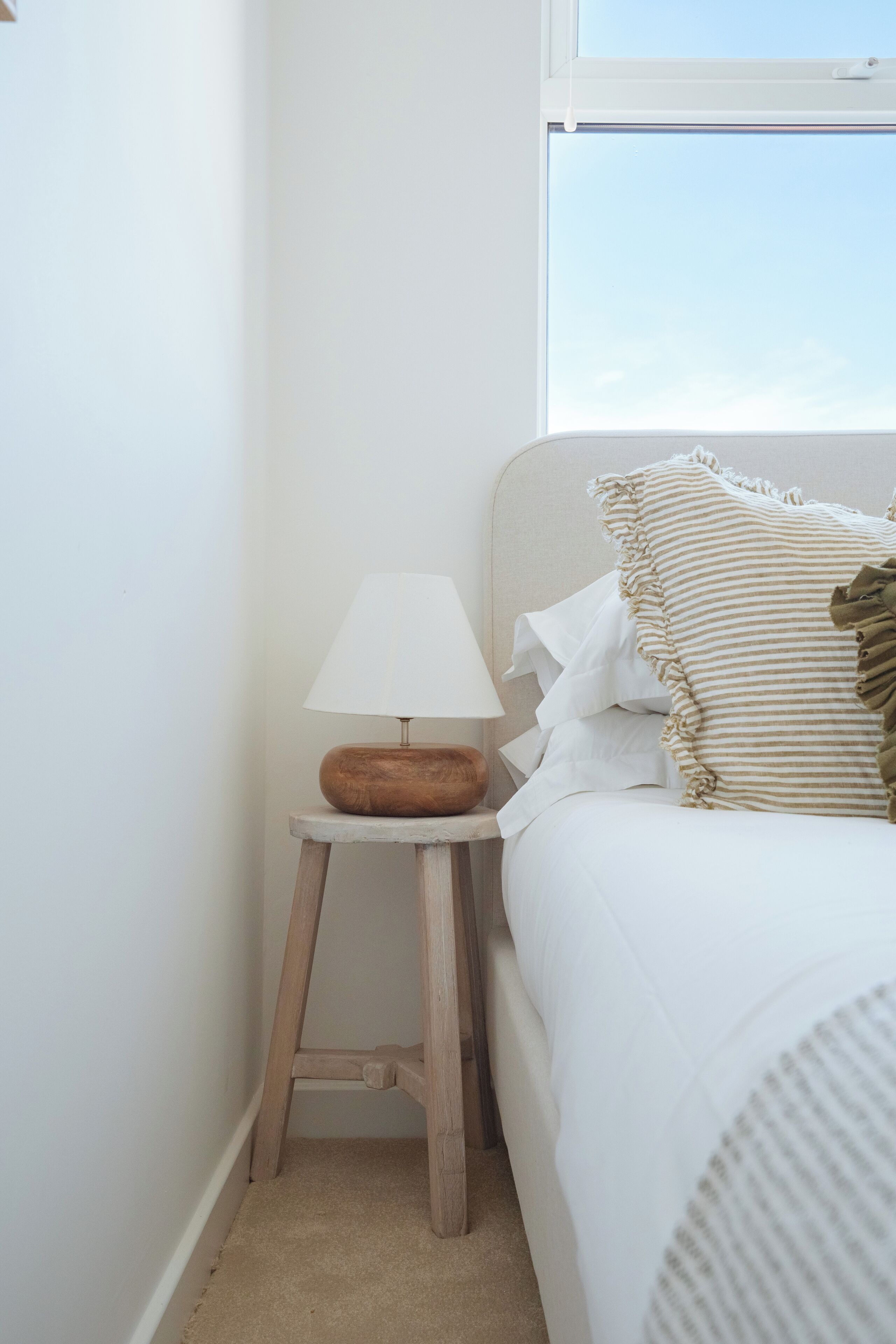 A neatly made bed with white bedding and a striped pillow, a small wooden stool with a white lamp, and a window with a view of the sky.