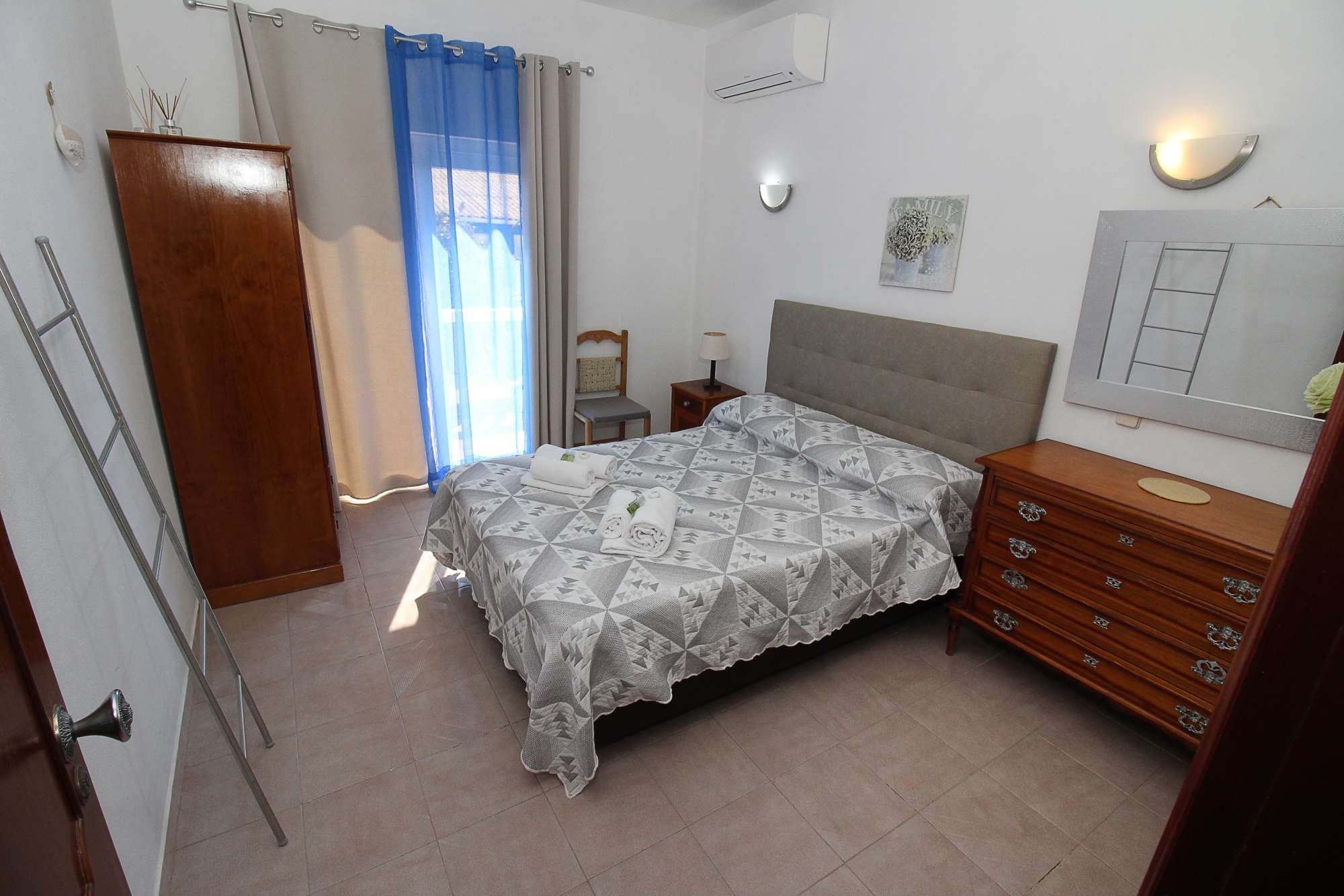 A bedroom with a bed, a dresser, a nightstand, a lamp, a mirror, and a window with curtains.