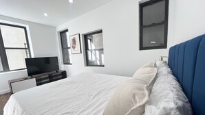 A modern bedroom with a large bed, a flat-screen TV, and a window with blinds. - 4-bedroom apartment in the lower east side (New York)