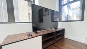 A modern living room with a flat-screen TV on a wooden stand, a remote control on the stand, and a large window providing natural light. - 4-bedroom apartment in the lower east side (New York)