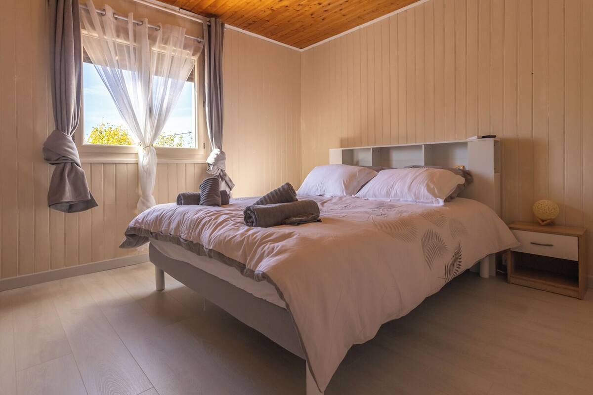 Een slaapkamer met een bed, een nachtkastje, een raam met gordijnen en een houten plafond.