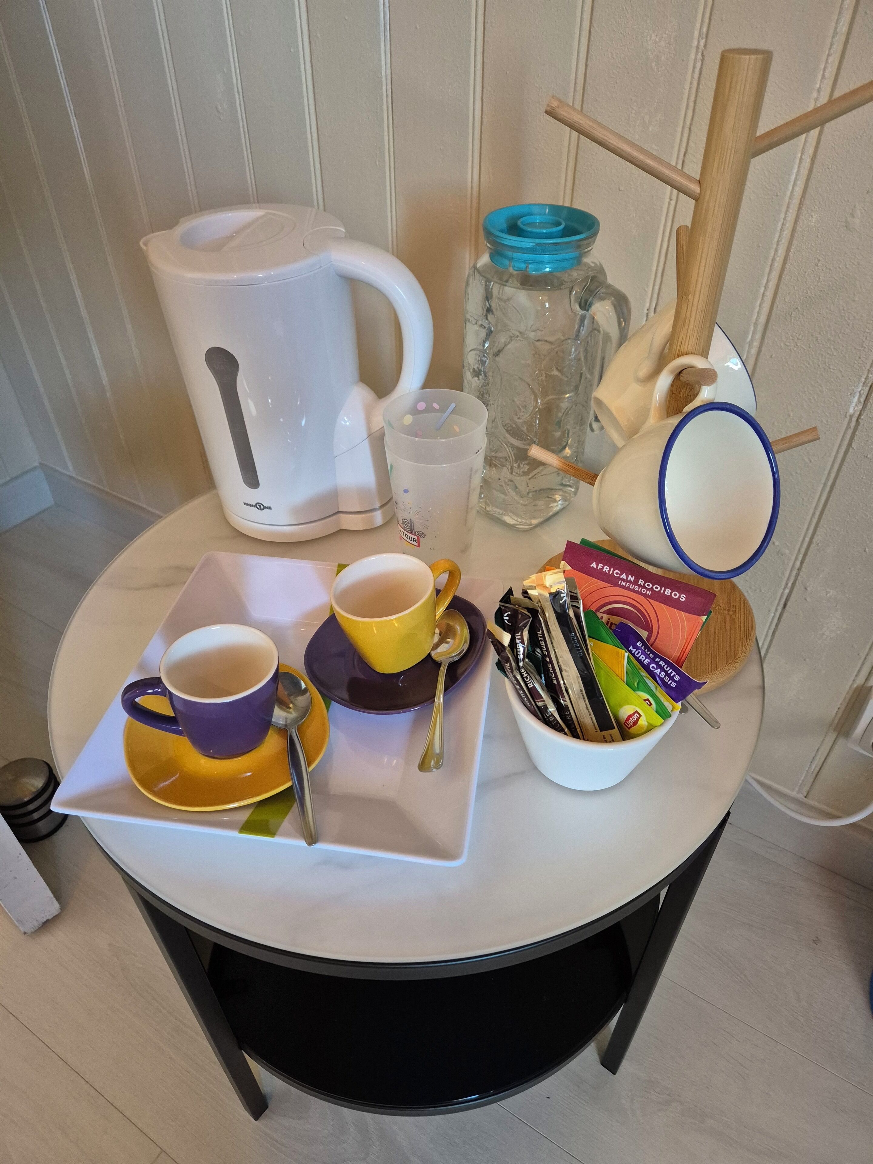 Una mesa con un hervidor blanco, dos tazas, un vaso, una cuchara de madera y un cuenco de aperitivos.