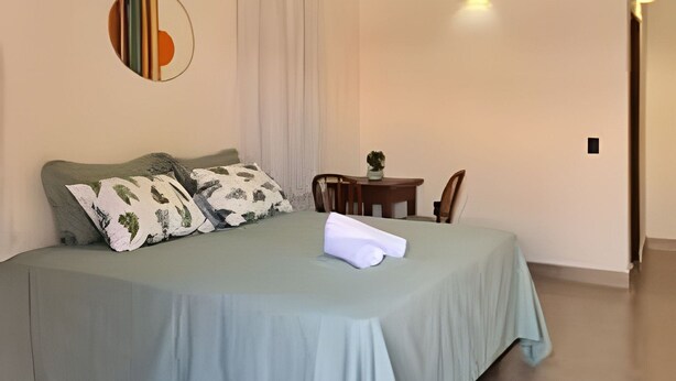 A neatly made bed with a light green bedspread and decorative pillows, a hanging light fixture, and a round mirror on the wall. - Chalet 800 meters from Chapada dos Veadeiros Park, Wi-Fi (Alto Paraíso de Goiás)