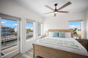 Une chambre à coucher comprenant un lit, un ventilateur de plafond, une table de chevet, une lampe et de grandes fenêtres donnant sur un quai et des bâtiments.