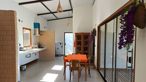 Un comedor con mesa y sillas, una cocina al fondo y un ventanal grande con plantas colgantes.
