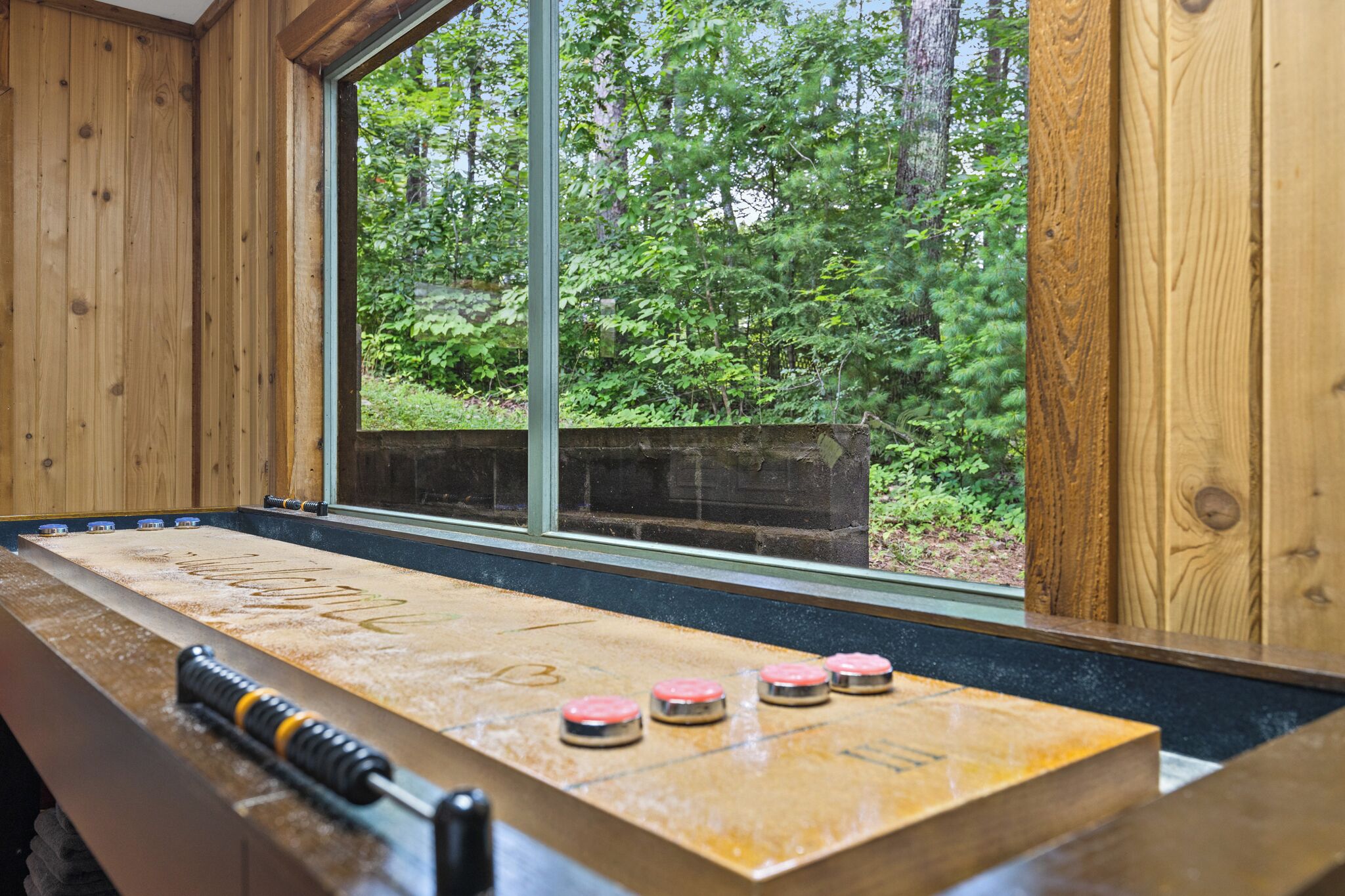 A shuffleboard table with pucks in a wooden room with large windows overlooking a forest.