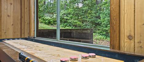 A shuffleboard table with pucks in a wooden room with large windows overlooking a forest.
