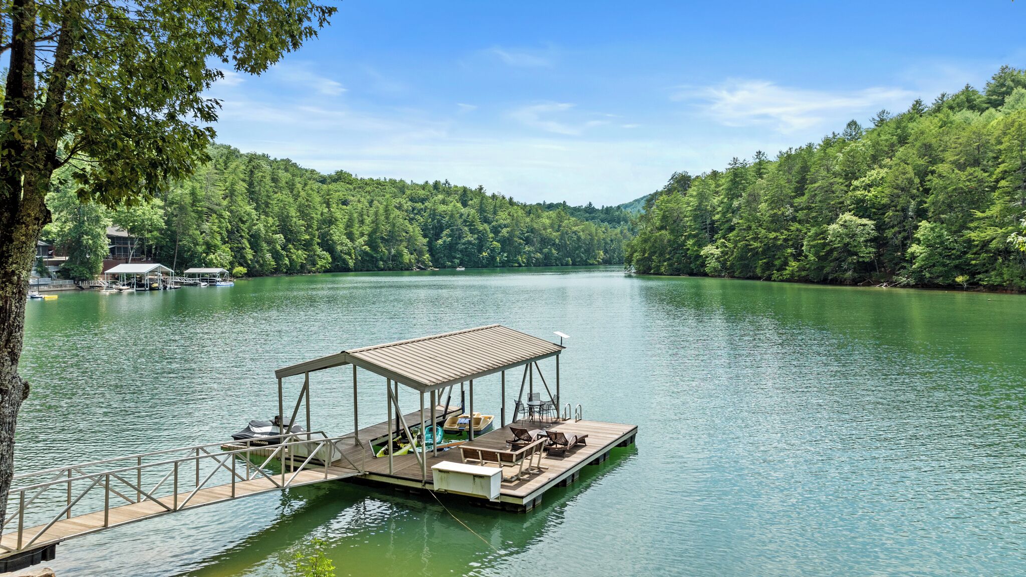 A docked boat with a covered seating area and a small building, surrounded by a forested lake.