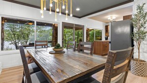 Un coin repas avec une table en bois, des chaises et un lustre. On aperçoit un réfrigérateur et une plante au fond.