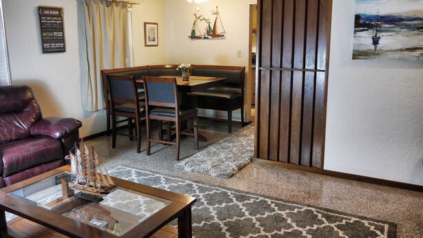 A living room with a leather sofa, a glass coffee table, a dining area with a table and chairs, a decorative sailboat wall hanging, and a painting of a seascape.