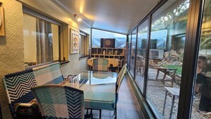 A covered patio area with a glass table and chairs, a bookshelf, and a view of the outdoor garden. - VELO APPARTEMENT BABY FOOT 3 VRAIES CHAMBRES 1 MINUTES DE LA MER JARDIN VERANDA (Nice)