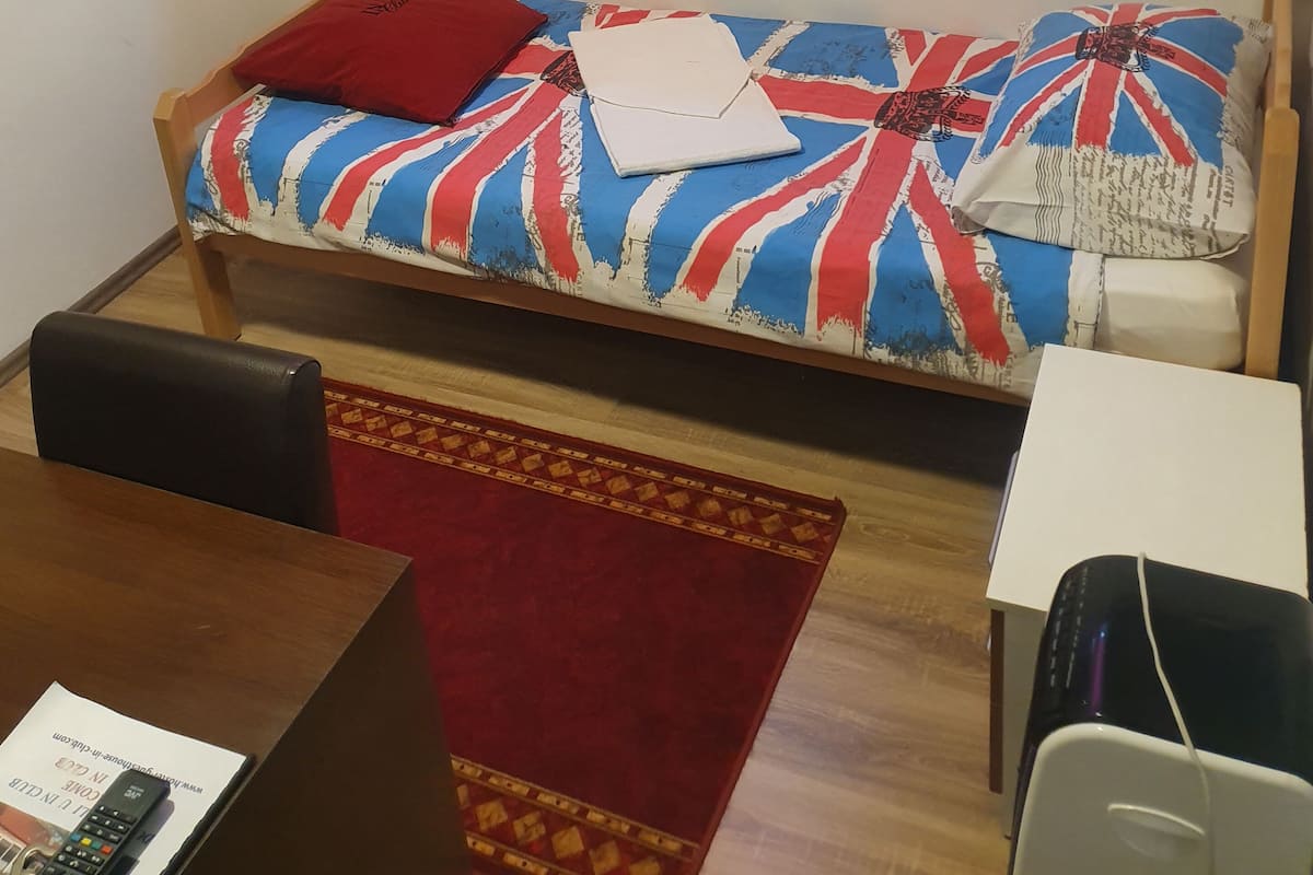 Ein kleines Zimmer mit Bett, Schreibtisch, Stuhl, Fernseher und Deckenleuchte.