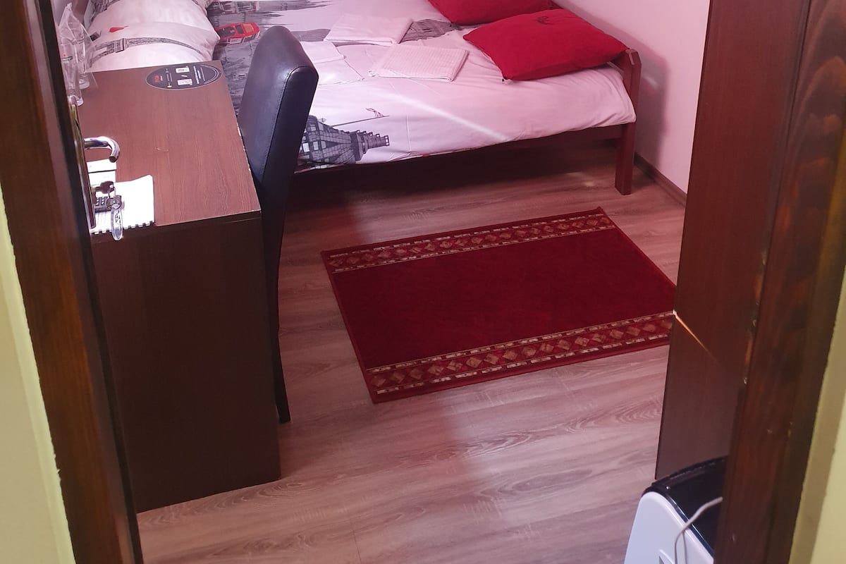 Ein kleiner, kompakter Raum mit einem Bett, einem Schreibtisch und einem Stuhl. Ein Fernseher ist an der Wand über dem Bett angebracht.