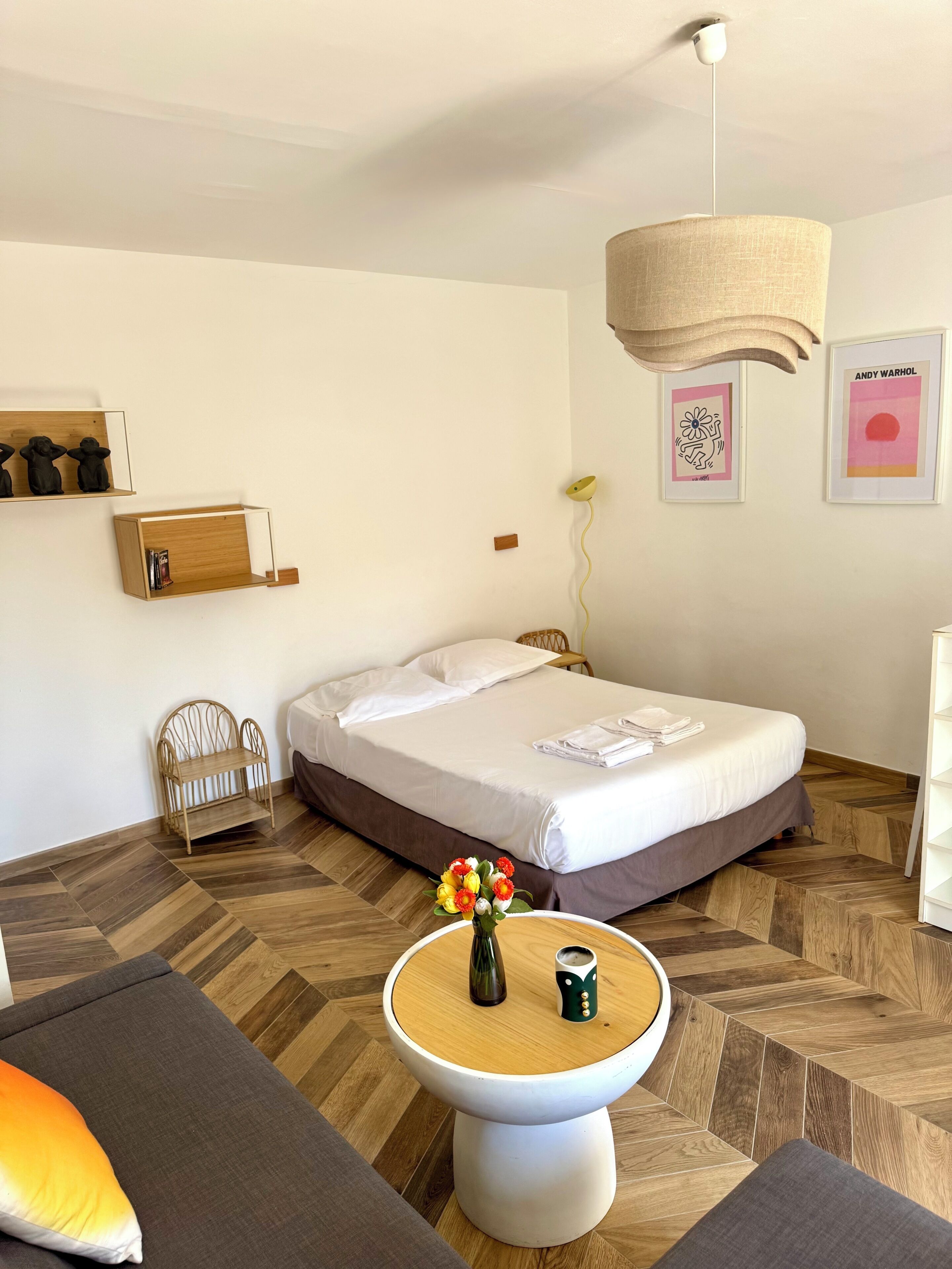 Hotelkamer met een bed, nachtkastje met een vaas en beker, een stoel, houten vloer en een wand met kunstwerken.