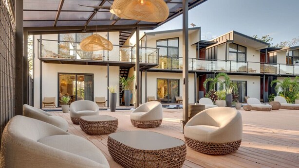 Un espacio moderno para sentarse al aire libre con mobiliario de mimbre y una estructura con paredes de vidrio.
