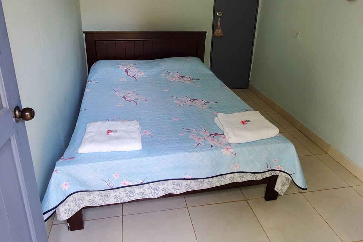 Una habitación pequeña con una cama individual, colcha azul estampada con flores, cabecero de madera y piso de baldosas.